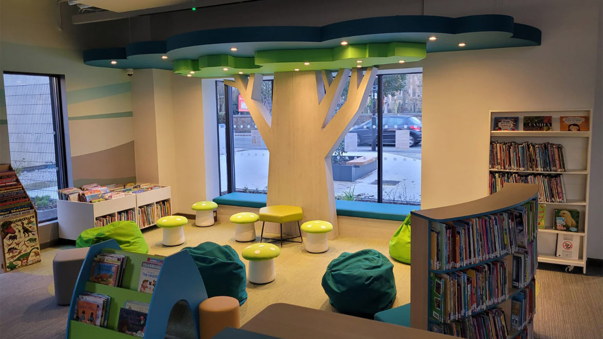 Little Lever Library modern interior children's area with small green stools and large tree wooden design in front of a large light window