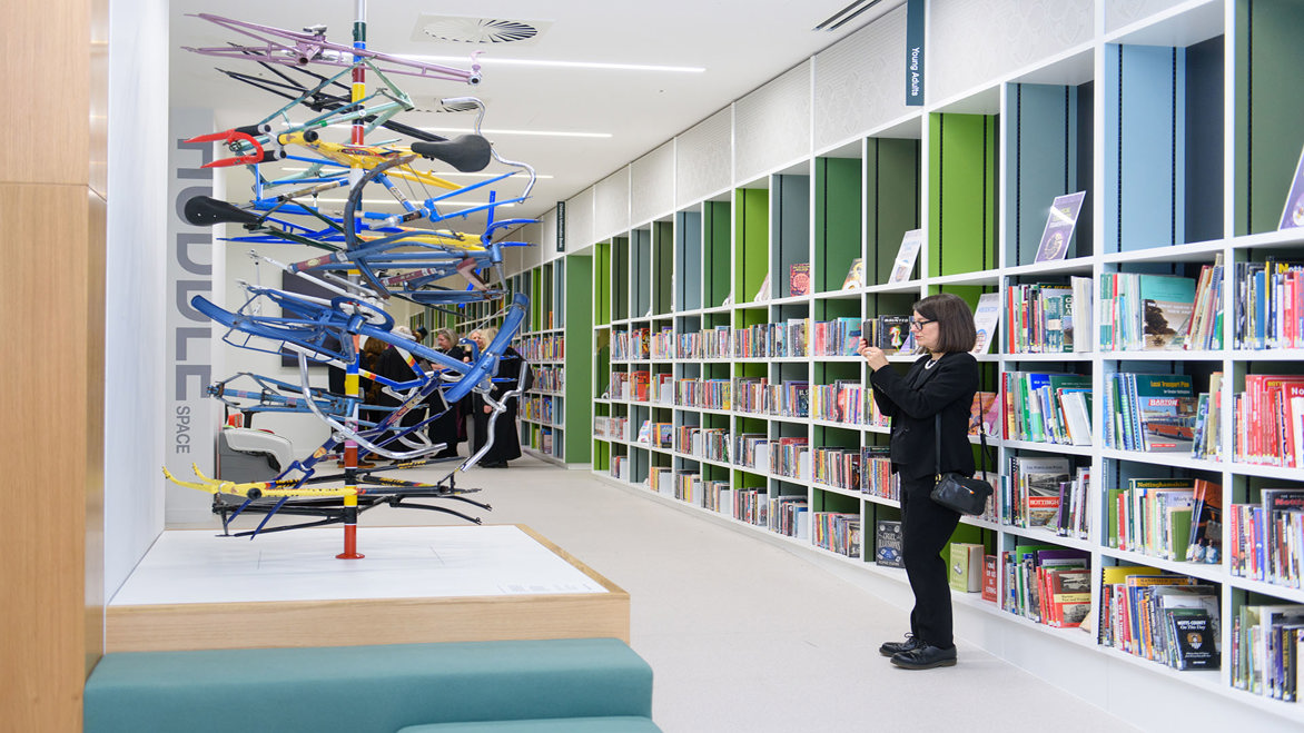 A long line of bookshelves and sculpture inside Nottingham Central Library, with a visitor taking a photograph