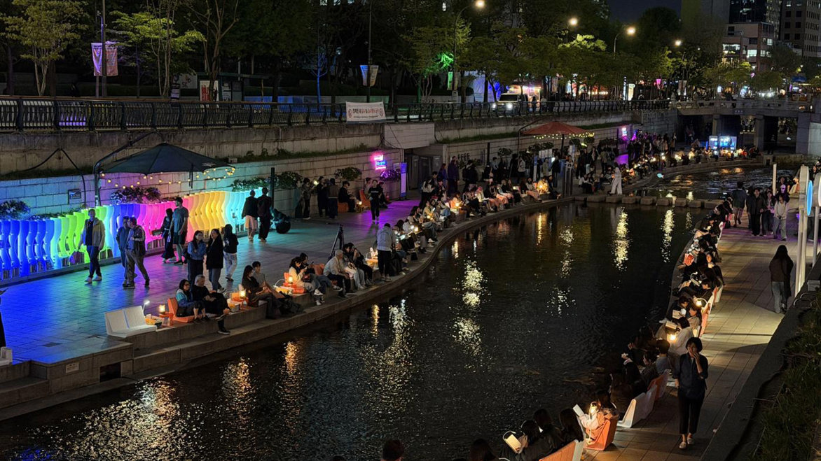 Evening photo of the outdoor library showing people sitting next to a river