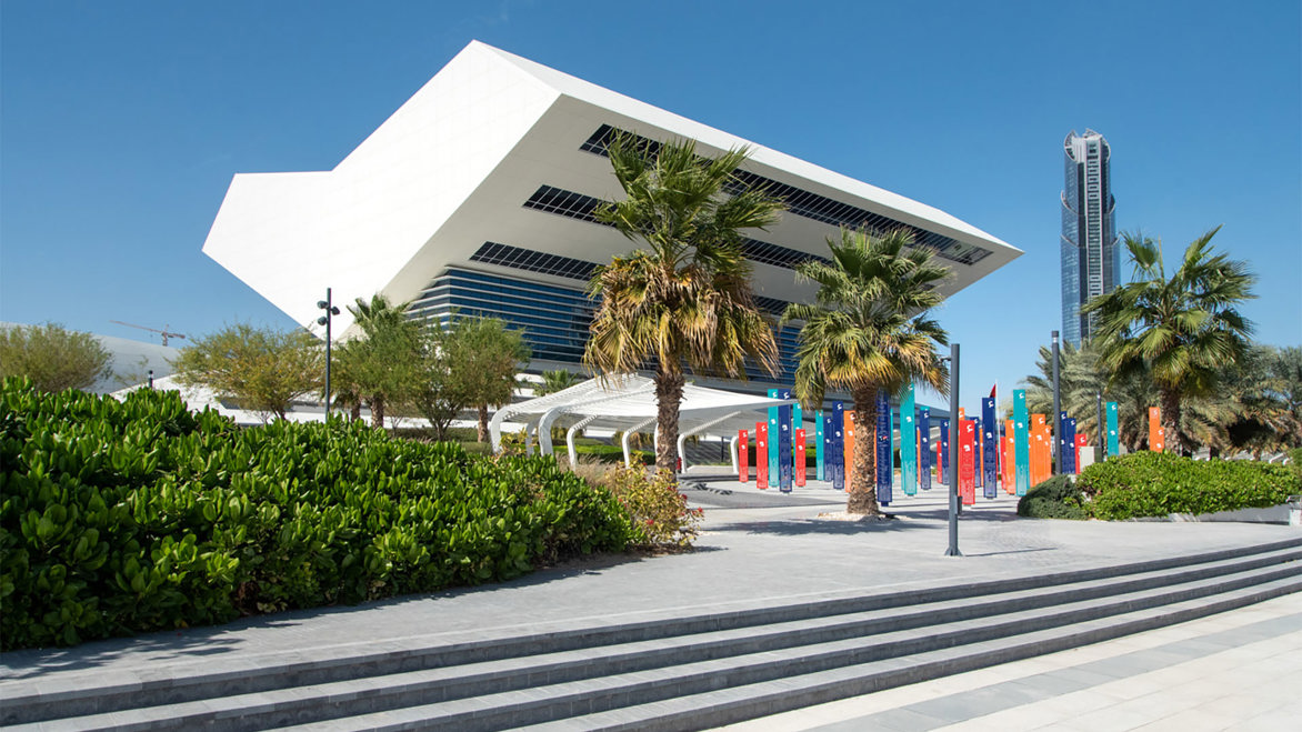 Exterior photograph of The Mohammed Bin Rashid Library in glorious sunshine with an array of palm trees surrounding the white building and steps leading up to it!