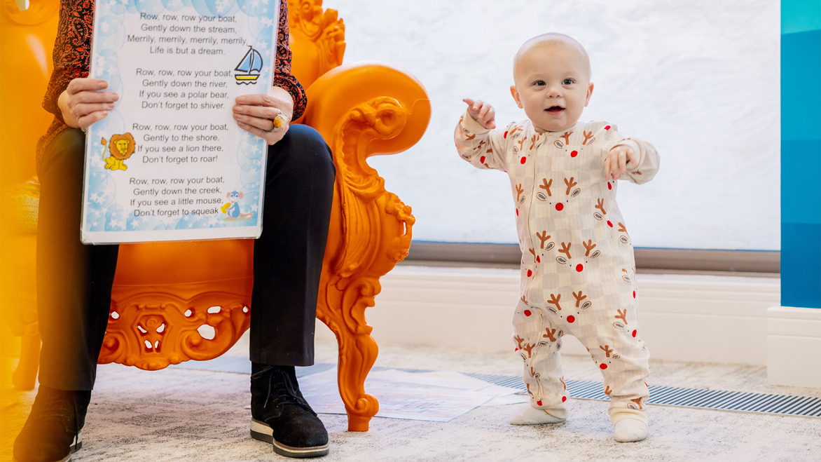 Cute baby standing up in St Helier children's library next to its mother reading a large book on the cosy orange chair